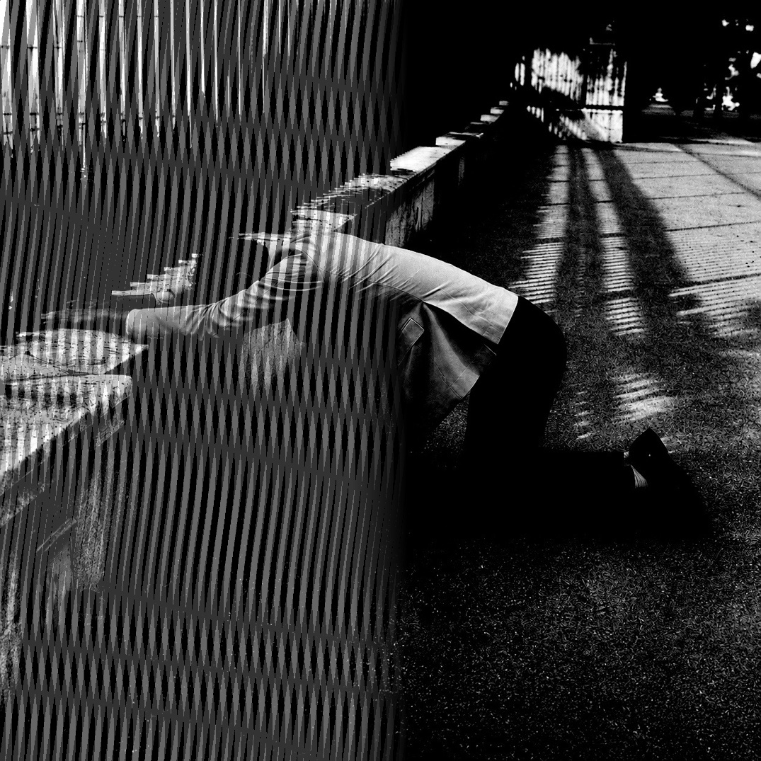 A figure kneeling beside a low wall, half the frame overlaid with the striped shadow of an iron fence, in black and white.