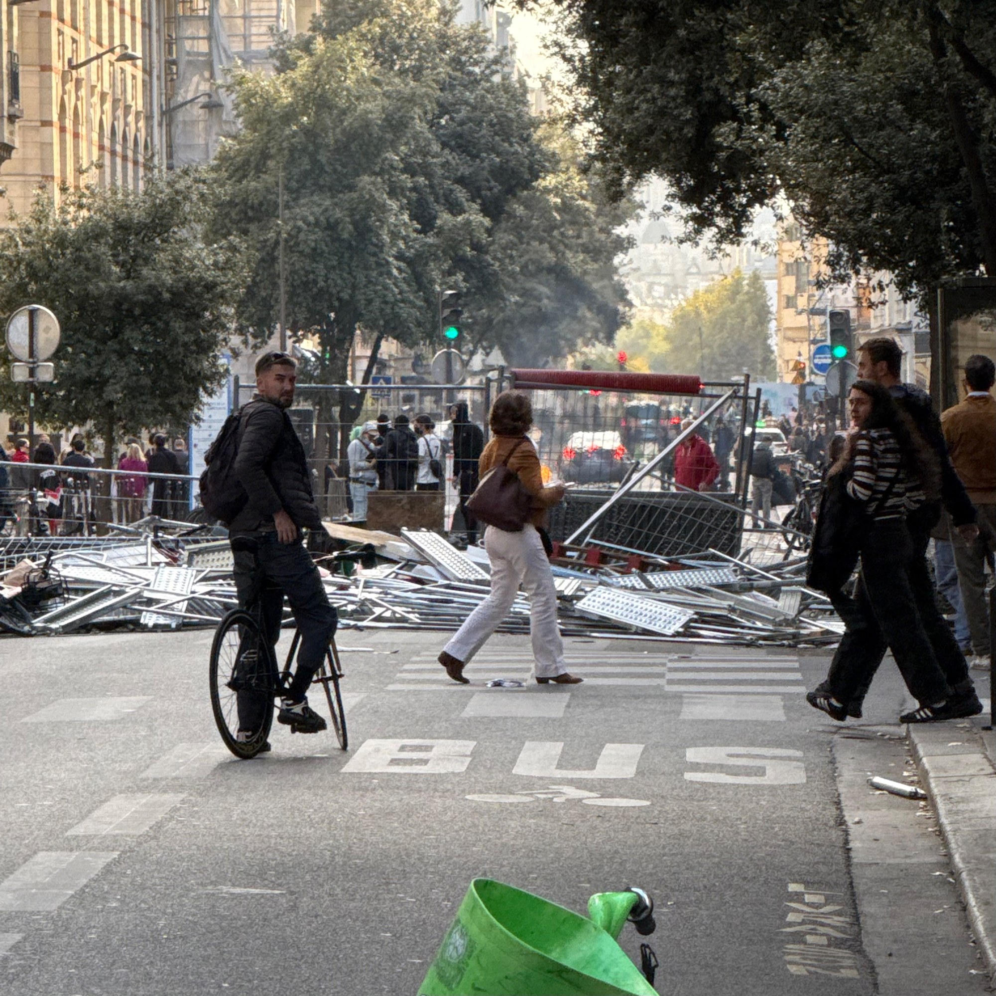 A city street blocked by a heap of fallen metal fencing and scaffolding. A man on a bicycle looks back over his shoulder as pedestrians cross in front of the pile; a green bike-share bin sits in the foreground.